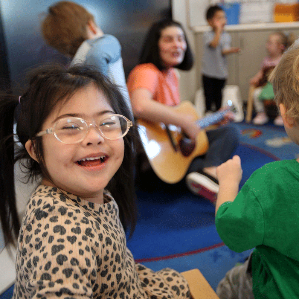 A girl with physical disabilities or developmental delays smiling with a group of children listening to the teacher playing guitar in a classroom - Rise School San Antonio